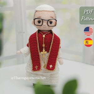 May include: A crocheted doll of a religious figure wearing a white robe, red stole, and gold cross. The doll has glasses and a white hat. The image includes the text "PDF Pattern" and the American and Spanish flags.