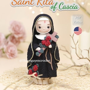 May include: A crochet doll of Saint Rita of Cascia wearing a black habit with gold trim, a white wimple, and a red mark on the forehead. The doll holds a brown wooden cross adorned with small red crocheted roses. Text reads Saint Rita of Cascia.