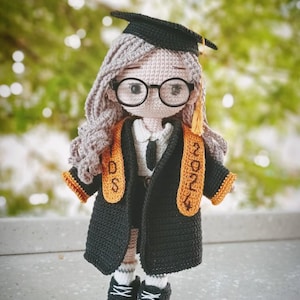 Puede incluir: Muñeca de ganchillo hecha a mano vestida con toga y birrete de graduación. La muñeca tiene pelo gris, gafas redondas y un birrete negro. La toga negra tiene detalles en naranja y una banda que dice "2024". La muñeca lleva zapatillas negras.