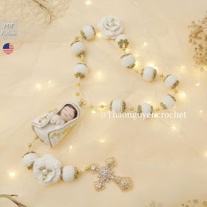 Può includere: Rosario all'uncinetto fatto a mano con perline bianche e dorate, un ciondolo a forma di croce e una piccola statuina di bambino. Il rosario presenta un motivo floreale e dettagli dorati. Il testo "PDF Pattern" e la bandiera americana sono inclusi.