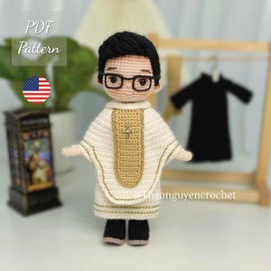 Puede incluir: Muñeco de ganchillo de un sacerdote con vestiduras blancas y doradas, zapatos negros y gafas. El muñeco tiene pelo negro y está de pie frente a un perchero de madera con una prenda negra. El texto "PDF Pattern" y una bandera estadounidense son visibles.