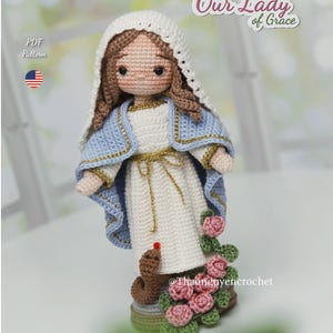 Op de afbeelding: Gehaakte pop van Onze-Lieve-Vrouw van Genade, gekleed in een witte jurk en een blauwe sjaal met gouden details. De pop heeft bruin haar en een witte sluier. Een slang en roze rozen staan aan de basis. De tekst "Our Lady of Grace" is zichtbaar.