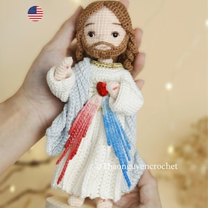 Puede incluir: Figura de Jesús tejida a ganchillo, con cabello y barba castaños, vistiendo una túnica blanca y un chal azul claro. Presenta un corazón rojo y rayos de luz rojos y azules. La figura está sobre una base de madera. Texto: PDF Pattern.