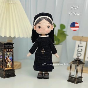 May include: A handmade crochet doll of a nun wearing a black habit, a white wimple, and a cross necklace. The doll has a detailed face and is standing upright. The image includes the text "PDF Pattern" and the American flag.