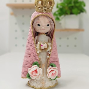 Puede incluir: Muñeca de ganchillo hecha a mano de una figura religiosa, con una capa rosa y una corona dorada. La muñeca tiene un vestido blanco, un collar de perlas y dos rosas rosas en la base. La muñeca mide aproximadamente 18 cm de altura.