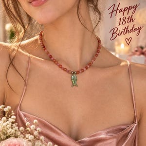 May include: A beaded necklace featuring small, round, reddish-orange stones and a single green, fish-shaped pendant. The text "Happy 18th Birthday" with a heart symbol is visible in the upper right corner of the image.