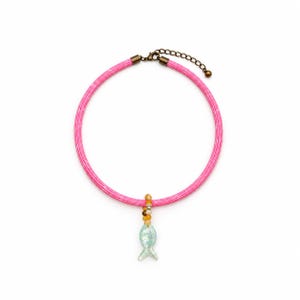 May include: A bright pink cord necklace featuring a light blue fish-shaped pendant suspended from a small cluster of yellow and orange beads. The necklace includes a bronze-toned metal clasp and an adjustable chain link extension for length.