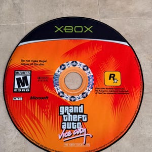 Puede incluir: Un disco negro de Xbox con un dise&ntilde;o naranja y rojo. El disco tiene el texto "Grand Theft Auto Vice City" y la clasificaci&oacute;n ESRB "M" para mayores de 17 a&ntilde;os.