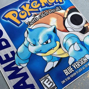 Puede incluir: Una alfombra de juego Game Boy Pokemon azul y amarilla con un personaje Pokemon azul y amarillo con el texto "Gotta catch 'em all!" y "Blue Version". La alfombra tambi&eacute;n incluye una clasificaci&oacute;n ESRB de "Everyone" y un temporizador que muestra "02:21:02".