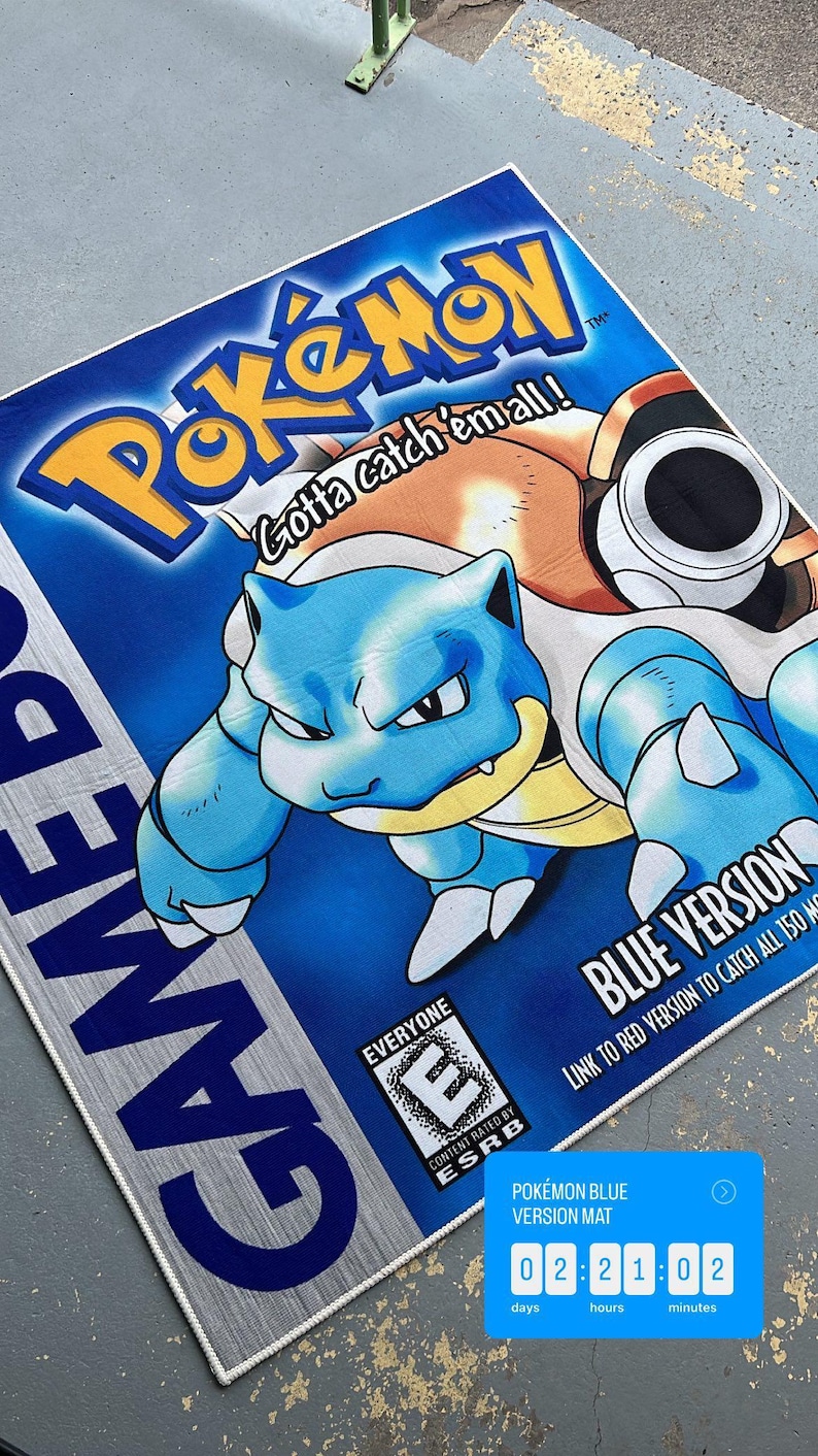 Puede incluir: Una alfombra de juego Game Boy Pokemon azul y amarilla con un personaje Pokemon azul y amarillo con el texto "Gotta catch 'em all!" y "Blue Version". La alfombra tambi&eacute;n incluye una clasificaci&oacute;n ESRB de "Everyone" y un temporizador que muestra "02:21:02".