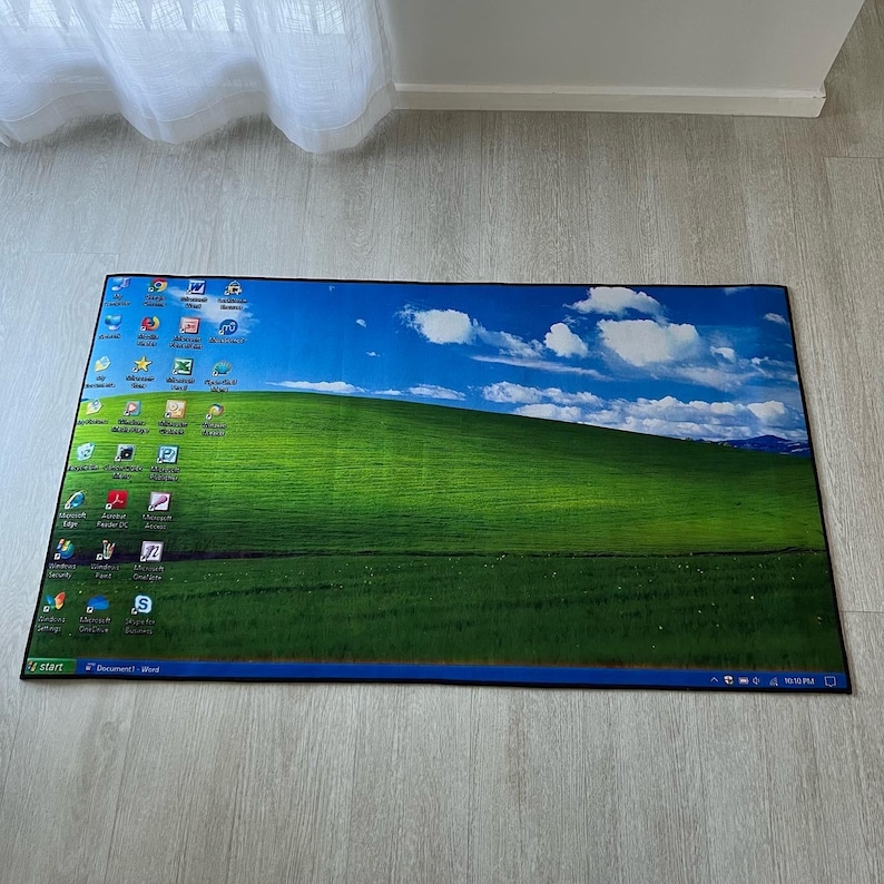 Puede incluir: Una alfombra de bienvenida azul y verde con una imagen del cl&aacute;sico fondo de escritorio de Windows XP. El fondo presenta un cielo azul con nubes blancas y una colina verde cubierta de hierba. Los iconos del escritorio son visibles en la esquina superior izquierda de la imagen. La esquina inferior derecha de la alfombra muestra la hora 10:15 PM.