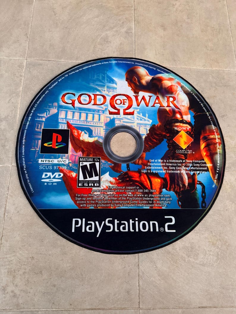Puede incluir: Un disco de juego de PlayStation 2 para God of War II. El disco es negro con un logotipo de PlayStation 2 plateado y el t&iacute;tulo del juego en blanco. El disco presenta un fondo rojo y naranja con un personaje del juego, Kratos, en el centro.