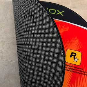 Puede incluir: Una alfombra redonda negra y roja con el logotipo de Rockstar Games y la palabra "Xbox" impresa en ella. La alfombra tiene un respaldo antideslizante negro.