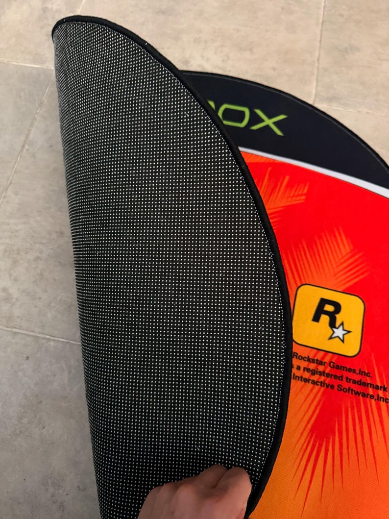 Puede incluir: Una alfombra redonda negra y roja con el logotipo de Rockstar Games y la palabra "Xbox" impresa en ella. La alfombra tiene un respaldo antideslizante negro.