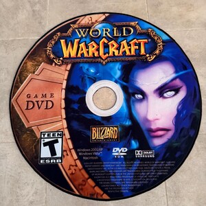 Puede incluir: Un DVD de juego World of Warcraft con un dise&ntilde;o azul y dorado que presenta a un personaje femenino con ojos blancos. El disco est&aacute; etiquetado como "Game DVD" y tiene una clasificaci&oacute;n Teen de la ESRB. La parte posterior del disco enumera los requisitos del sistema del juego y la informaci&oacute;n de copyright.