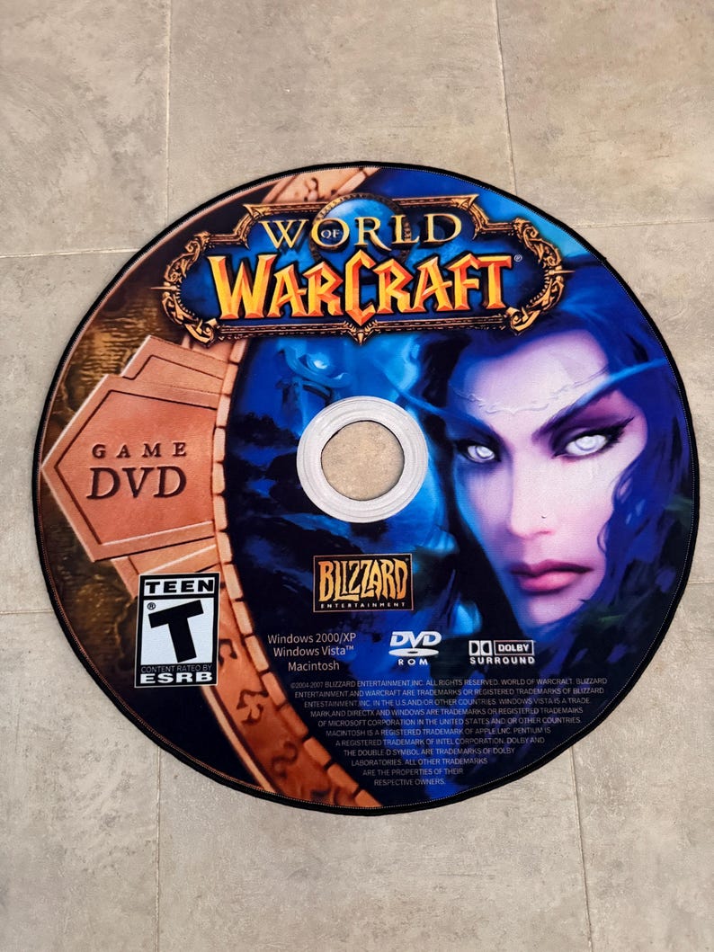 Puede incluir: Un DVD de juego World of Warcraft con un dise&ntilde;o azul y dorado que presenta a un personaje femenino con ojos blancos. El disco est&aacute; etiquetado como "Game DVD" y tiene una clasificaci&oacute;n Teen de la ESRB. La parte posterior del disco enumera los requisitos del sistema del juego y la informaci&oacute;n de copyright.