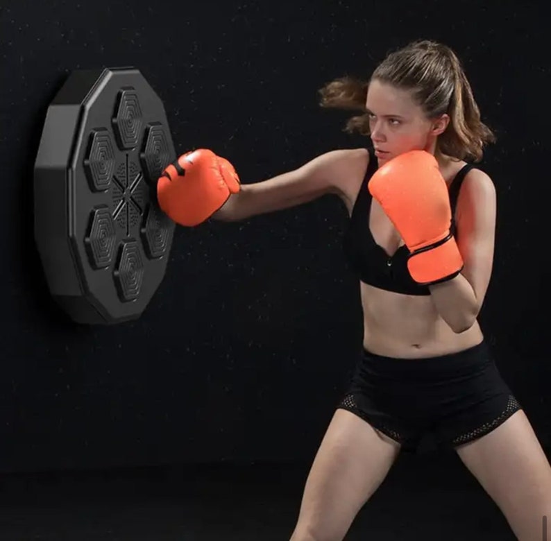May include: A woman in orange boxing gloves and black sportswear practices boxing with a black, hexagonal, wall-mounted punching target. The target has six hexagonal pads and a central star design.