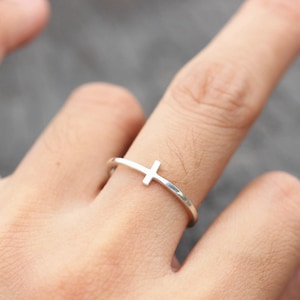 Könnte beinhalten: Ein silberner Ring mit einem schlichten Kreuzdesign. Der Ring ist dünn und zart, das Kreuz ist mittig auf dem Band platziert.