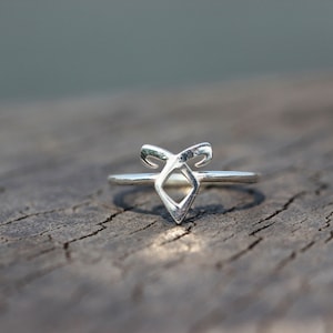 Könnte beinhalten: Ein silberner Ring mit einem stilisierten Design, das eine rautenförmige Form in der Mitte aufweist, flankiert von gebogenen Elementen. Der Ring liegt auf einer strukturierten Holzoberfläche mit unscharfem Hintergrund.