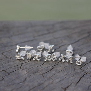May include: A set of silver initial earrings with a cursive font. The earrings are displayed on a wooden surface.
