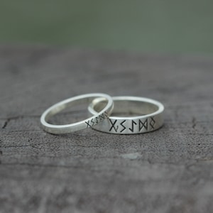 Könnte beinhalten: Zwei silberne Ringe mit gravierten Runen. Die Ringe liegen übereinander.