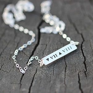 Puede incluir: Un collar de plata con un colgante de barra grabado con los números romanos VII y VIII separados por un triángulo.