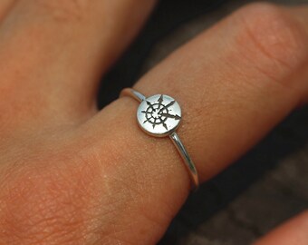 May include: A silver ring with a circular design featuring a compass rose. The ring is worn on a finger.