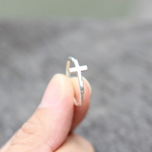 May include: A silver ring featuring a simple cross design. The ring is thin and delicate, and the cross is small and understated.