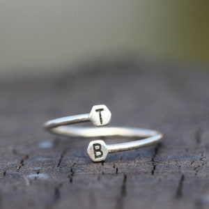 Könnte beinhalten: Ein silberner Ring mit zwei sechseckigen Anhängern, einer mit dem Buchstaben "T" und der andere mit dem Buchstaben "B".