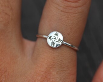 May include: A silver ring with a circular design featuring a geometric pattern. The ring is worn on a finger.