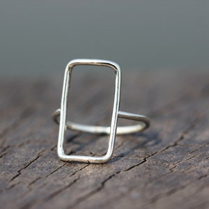 Könnte beinhalten: Ein silberner Ring mit einem rechteckigen, offenen Design. Der Ring hat ein dünnes Band und eine geometrische, offene Rechteckform. Der Ring wird auf einer Holzoberfläche gezeigt.