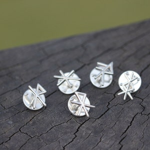 May include: Five silver pins with different geometric designs. The pins are arranged in a semi-circle on a wooden surface.