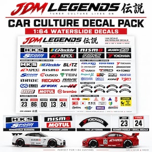 May include: A 1:64 scale waterslide decal pack featuring various automotive brand logos, tire lettering, and number panels. The sheet includes text such as JDM LEGENDS, HKS, GReddy, Nismo, and Mugen, displayed alongside two model cars with applied decals.