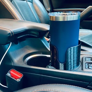 May include: A navy blue insulated tumbler with a silver rim and clear lid sits in a black 3D-printed car cup holder. The cup holder is placed in a car's center console, next to a seatbelt buckle.