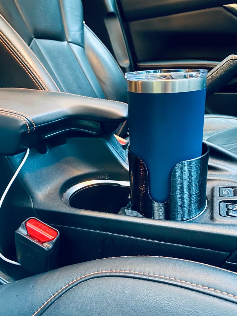 May include: A navy blue insulated tumbler with a silver rim and clear lid sits in a black 3D-printed car cup holder. The cup holder is placed in a car's center console, next to a seatbelt buckle.