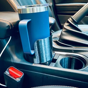 May include: A blue insulated tumbler with a handle sits in a black 3D-printed car cup holder. The cup holder is placed in a car's center console. A red seatbelt buckle is visible.