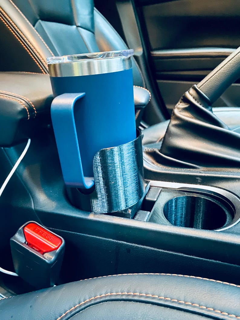 May include: A blue insulated tumbler with a handle sits in a black 3D-printed car cup holder. The cup holder is placed in a car's center console. A red seatbelt buckle is visible.