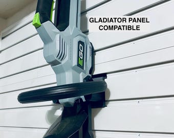May include: A gray and black EGO 56V power tool mounted on a white wall panel. The tool features a green trigger and green branding. A white label with black text reads GLADIATOR PANEL COMPATIBLE. The mount is black and holds the tool securely.