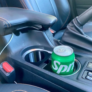 May include: A green Sprite soda can in a car's cup holder. The can has white text and is surrounded by a black interior. The car seat is black with orange stitching.