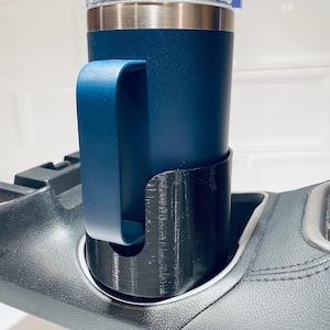 May include: A navy blue insulated travel mug with a handle, placed in a black cup holder. The mug has a silver rim and a clear lid. The cup holder is designed to fit into a car's console.