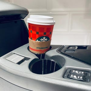 May include: A red Starbucks coffee cup with a white lid sits in a car's cup holder. The cup has a brown sleeve and a green and black plaid design. The Starbucks logo is visible on the cup.