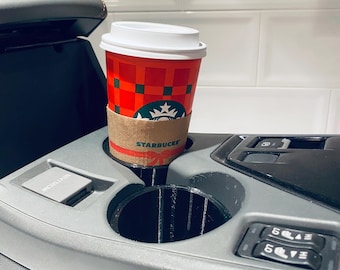 May include: A red Starbucks coffee cup with a white lid sits in a car's cup holder. The cup has a brown sleeve and a green and black plaid design. The Starbucks logo is visible on the cup.