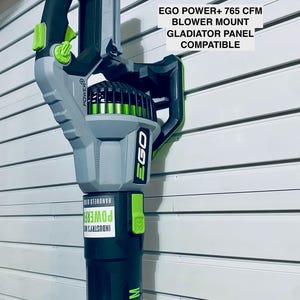 Puede incluir: Un soporte para soplador EGO Power+ 765 CFM, compatible con paneles Gladiator. El soplador de mano es principalmente gris y negro, con detalles en verde. El texto "EGO POWER+ 765 CFM BLOWER MOUNT GLADIATOR PANEL COMPATIBLE" es visible.