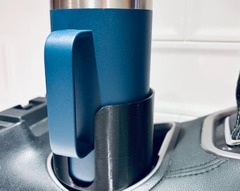 May include: A dark blue insulated tumbler with a matching handle sits inside a black cylindrical cup holder adapter. The tumbler features a stainless steel rim and a clear lid, while the adapter is secured within a vehicle center console.