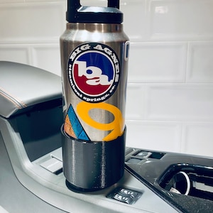 May include: A stainless steel water bottle with a black handle and a "Big Agnes" sticker, resting in a black cup holder. The bottle has a black lid and is cylindrical. The cup holder is in a car's console.