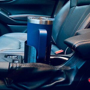 May include: A navy blue insulated tumbler with a handle sits in a black car cup holder. The tumbler has a silver rim and a clear lid. The cup holder is designed to fit the car's center console.