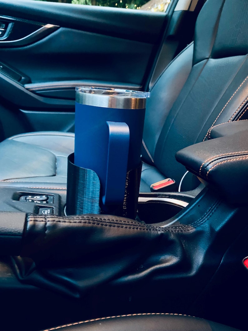 May include: A navy blue insulated tumbler with a handle sits in a black car cup holder. The tumbler has a silver rim and a clear lid. The cup holder is designed to fit the car's center console.
