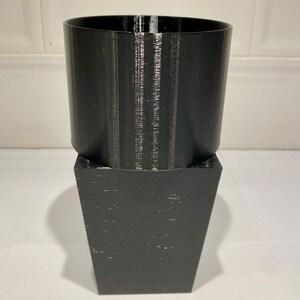 Peut inclure: Un pot de fleurs noir imprim&eacute; en 3D avec un dessus cylindrique et une base carr&eacute;e.