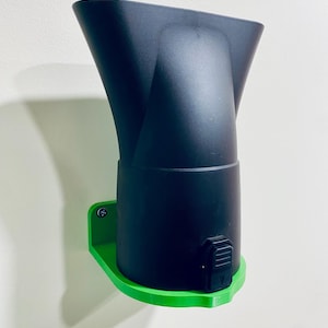 May include: A black hair dryer attachment mounted on a bright green holder. The attachment has a wide, flared nozzle and a cylindrical base. The holder is attached to a wall with a screw.
