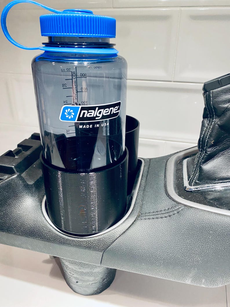 May include: A clear Nalgene water bottle with a blue lid and a black car cup holder. The bottle has measurement markings and the Nalgene logo. The cup holder is designed to fit in a car's center console.